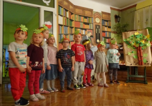 Dzieci w opaskach na głowie recytują wiersz. Obok stoi sztaluga z drzewem.