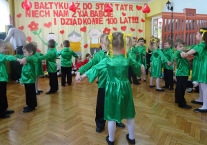 W kole dziewczynki w zielonych sukienkach oraz chłopcy w zielonych koszulach i czarnych spodniach wykonują taniec dla babci i dziadka