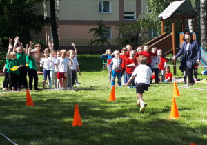 Przedszkolaki stoją w tzrech rzędach. Jeden chłopiec biegnie pomiędzy pachołkami.