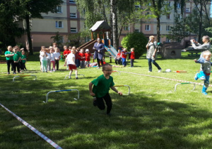 Przedszkolaki stoją w tzrech rzędach. Troje dzieci biegnie między przeszkodami.