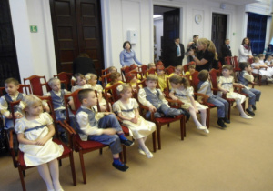 Przedszkolaki siedzą na krzesełkach w dużej sali obrad.