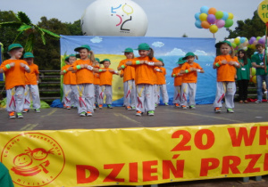 Przedszkolaki w pomarańczowych podkoszulkach, szarych spodniach i zielonych czapkach z daszkiem wykonują taniec hip-hop. W tle balony oraz tabliczka z logo przedszkola..