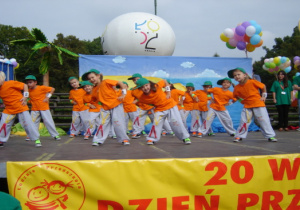 Przedszkolaki w pomarańczowych podkoszulkach, szarych spodniach i zielonych czapkach z daszkiem wykonują taniec hip-hop. W tle balony.
