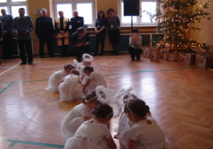 Dziewczynki w białych sukienkach kucają w tańcu. Policjanci oraz inni zebrani goście podziwiają występ dzieci.