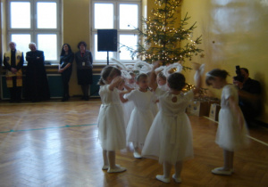Dzieci w białych sukienkach wykonują taniec z rękoma uniesionymi do góry. Policjanci oraz inni zebrani goście podziwiają występ dzieci.