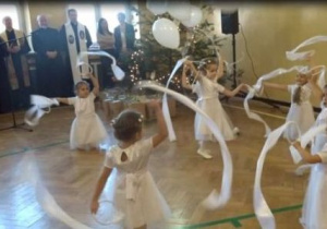 Dzziewczynki w białych sukienkach tańczą z szarfami. jedna dziewczynka trzyma białe balony. Zebrani goście podziwiają występ dzieci.