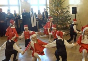 Dzieci w strojach mikołajowych tańczą w kole. Zebrani goście podziwiają występ dzieci.