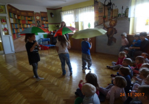 Troje dzieci z parasolami oraz prowadząca. Pozostałe dzieci siedzą i oglądają występy.