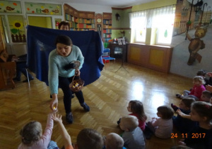 Prowadząca z koszykiem w ręku rozdaje dzieciom światełak. Za nią druga prowadząca trzyma niebieski materiał.