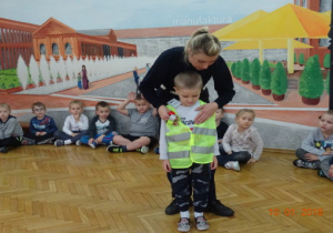 Dzieci siedzą, policjantka zakładaz chłopcu kamizelkę odblaskową.