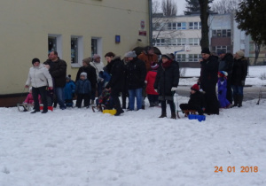 Dzieci i rodzice stoją w trzech rzędach. W każdym rzędzie dziecko siedzi na sankach.