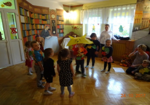 Dzieci stoją w półkolu, na ubraniach mają jesienne liście. W środku stoi chłopiec z parasolem. W tle nauczycielka