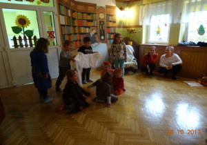 Czworo dzicei stoi trzymając biały materiał, czworo dzieci kuca. W tle dwie nauczycielki.
