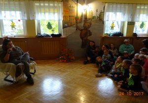 Na fotelu siedzi rodzic i czyta bajkę. Wszyscy z uwagą jej słuchają.