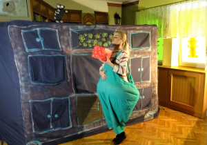Scenografia teatralna – kobieta przebrana za otyłą dziewczynkę stoi na tle szafy, w ręku trzyma pudełko, z którego dochodzi zapach smakołyków.