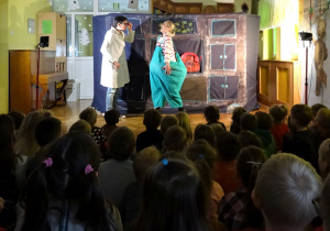 Sala gimnastyczna – scenografia teatralna, kobieta przebrana za otyłądziewczynkjest badana przez doktora, ma otwartą buzię i sprawdzane gardło. Dzieci siedzą i oglądają przedstawienie.