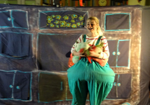 Scenografia teatralna – kobieta przebrana za otyłą dziewczynkę na tle szafy śpiewa piosenkę o zdrowym odżywianiu. W rękach trzyma zdrowe smakołyki – warzywo i owoc.