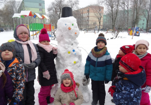 Zaśnieżony ogród przedszkolny – ósemka dzieci pozuje do zdjęcia z ogromnym bałwanem, złożonym z 3 kul, w czapce z wiadra, z nosem z marchewki, rękami z patyków oraz oczami i guzikami z kolorowych nakrętek.