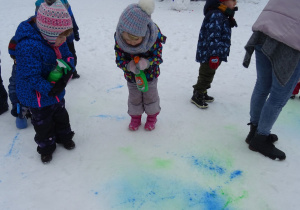 Zaśnieżony ogród przedszkolny – dwójka dzieci stoi na śniegu z farbami w sprayu w rękach – malują na śniegu. Obok nich nauczycielka i inne dziecko odwrócone tyłem.