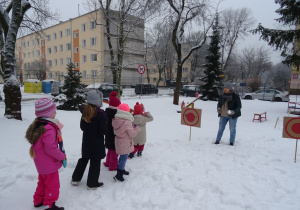 Zaśnieżony ogród przedszkolny – dzieci stoją w rzędzie, przed nimi czerwona tarcza, obok niej nauczycielka ze śnieżką w ręku tłumaczy zasady zabawy.