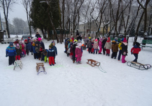Zaśnieżony ogród przedszkolny – cztery grupy przedszkolaków stoją w rzędach, przed każdą z nich sanki. Dwie nauczycielki nadzorują zabawę.