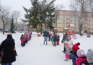 Zaśnieżony ogród przedszkolny – dwie grupy przedszkolaków stoją w rzędach, pozostałe dwie grupy w rzędach naprzeciwko. Dzieci przeciągają 2 liny ze wstążką zawiązaną pośrodku. Pomiędzy dwiema grupami przeciągającymi linę stoi czerwona tarcza do rzutu do celu. Cztery nauczycielki nadzorują zabawę.