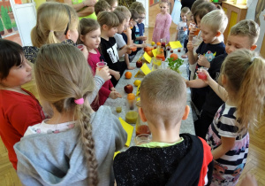 Sala gimnastyczna, na niej złączone stoliki, przykryte ceratkami a na nich kolorowe soki przygotowane do degustacji. Dookoła stolików grupa dzieci starszych testująca różne smaki soków.