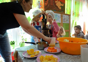 Dwa złączone stoliki, na nich stoi sokowirówka oraz miseczki z pomarańczami oraz marchewkami. Wokół stolików czworo dzieci oraz nauczycielka. Dwoje dzieci w fartuszkach i czepkach kuchennych obsługuje sokowirówkę, nauczycielka podstawia dzbanek, do którego spływa robiony przez dzieci sok.