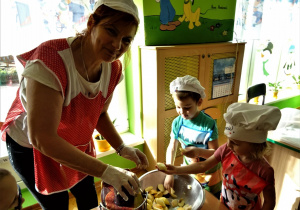 Stolik, na nim sokowirówka i miska z jabłkami. Przy nich pani kucharka i dwoje dzieci w strojach kuchennych. Pani kucharka obsługuje sokowirówkę, natomiast dzieci podają jej owoce.