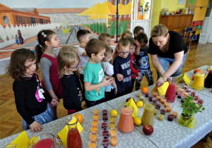 Sala gimnastyczna, na niej złączone stoliki, przykryte ceratkami a na nich kolorowe soki przygotowane do degustacji. Przy stolikach grupa dzieci i nauczycielka prezentująca soki.