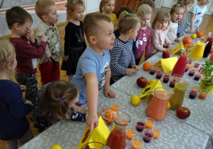 Sala gimnastyczna, na niej złączone stoliki, przykryte ceratkami a na nich kolorowe soki przygotowane do degustacji. Przy nich grupa dzieci jedno z nich – chłopiec pokazuje palcem sok, na który ma ochotę.