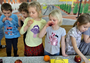 Pięcioro dzieci przy stole do degustacji soków. Czworo z nich próbuje kolorowych soków, jedno natomiast sięga po wybrany sok.