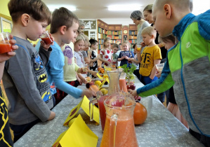 Sala gimnastyczna, na niej złączone stoliki, przykryte ceratkami a na nich kolorowe soki przygotowane do degustacji. Wzdłuż stolików grupa dzieci degustująca różne smaki soków. Degustację nadzorują dwie nauczycielki.