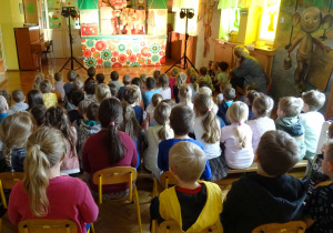 Scengrafia teatralna: kwiatki, drzewo oraz postać Szarego. 2 reflektory oświetlające scenę. Przedszkolaki siedzą i oglądają przedstawienie.