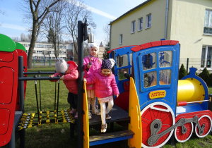 Lokomotywa a w niej bawiące się dzieci. Jedna dziewczynka schodzi po schodku, druga dziewczynka dzwoni dzwonkiem, trzecia przechodzi przez przejście do wagonu.