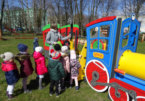 Dzieci z nauczycielką w ogrodzie przedszkolnym stoją z nauczycielką przy lokomotywie. Nauczycielka pociąga za dzwonek.