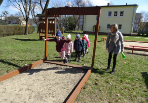 Ogród przedszkolny. Dzieci ustawione są w do wykonania skoku w dal. Jedno dziecko skacze. Nauczycielka nadzoruje zabawę.