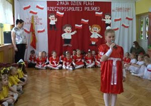 Na tle biało - czerwonej dekoracji na środku sali odświętnie ubrana dziewczynka recytuje wiersz. Po bokach siedzą przedszkolaki.