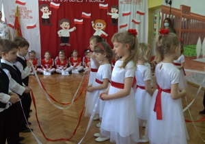 Na tle biało - czerwonej dekoracji odświętnie ubrane dzieci tańczą w parach z biało- czerwonymi wstążkami.