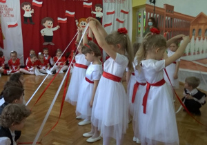 Na tle biało - czerwonej dekoracji odświętnie ubrane dzieci tańczą w parach z biało- czerwonymi wstążkami; chłopcy kucają.