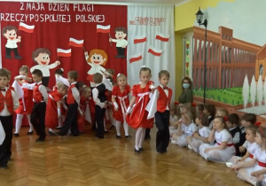 Na tle biało - czerwonej dekoracji odświętnie ubrane dzieci tańczą Poloneza. Z boku siedzą przedszkolaki z nauczycielką.