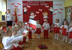 Na tle biało - czerwonej dekoracji, dzieci ubrane na biało - czerwono stoją w półkolu i recytują wiesz machając chorągiewkami.