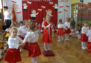 Na tle biało - czerwonej dekoracji odświętnie ubrane dzieci tańczą w parach wokół dziewczynki, która śpiewa piosenkę. Po bokach siedzą przedszkolaki.