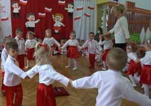 Na tle biało - czerwonej dekoracji odświętnie ubrane dzieci tańczą w kole z nauczycielką.