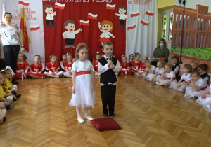 Na tle biało - czerwonej dekoracji dwoje odświętnie ubranych dzieci na środku sali recytuje wiersz. Po bokach siedzą przedszkolaki.