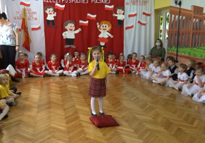 Na tle biało - czerwonej dekoracji odświętnie ubrana dziewczynka na środku sali recytuje wiersz. Po bokach siedzą przedszkolaki.