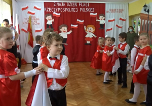 Na tle biało - czerwonej dekoracji odświętnie ubrane dzieci na środku sali tańczą w parach z biało - czerwonymi chustkami.
