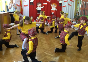 Na tle biało - czerwonej dekoracji odświętnie ubrane dzieci tańczą Polkę bałucką.