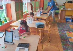 Dzieci pracują przy stolikach na taletach oraz na dywanie z robotami na planszach.
