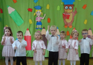 Dzieci stoją w półkolu na tle dekoracji i śpiewają piosenkę.
